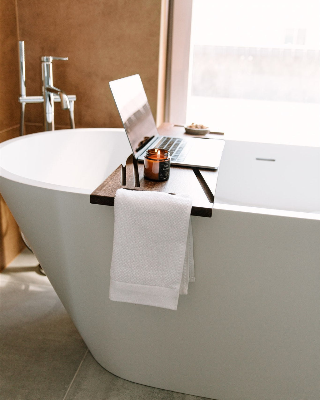 Laptop on a wooden tray over a bathtub with a cup and towel in a bathroom setting