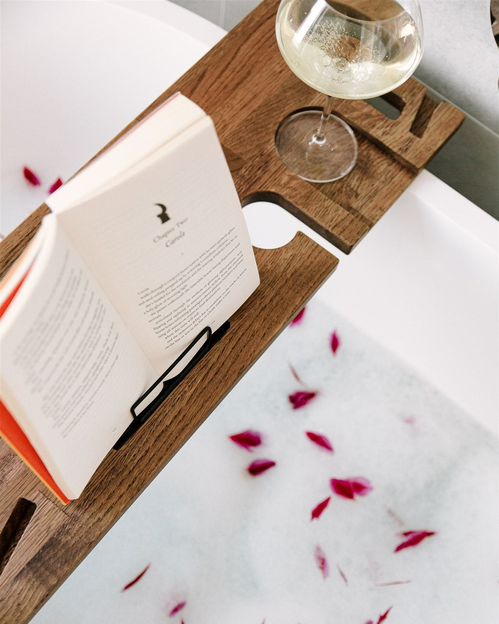 Open book on a wooden tray over a bathtub with rose petals and a glass of white wine.