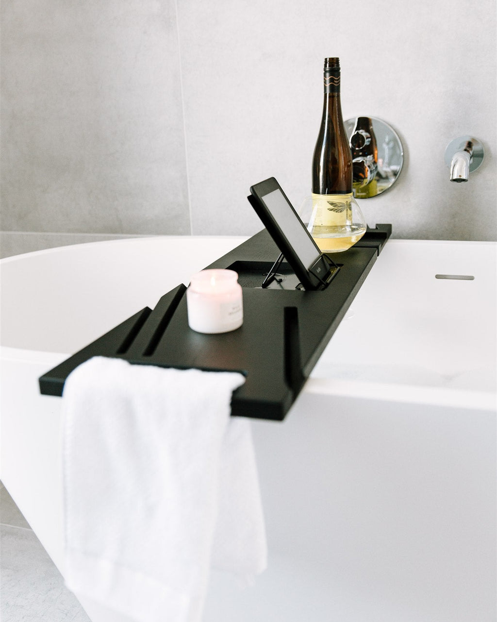 Black bathtub tray with a bottle, candle, and phone on a white bathtub.