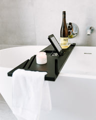 Black bathtub tray with a bottle, candle, and phone on a white bathtub.