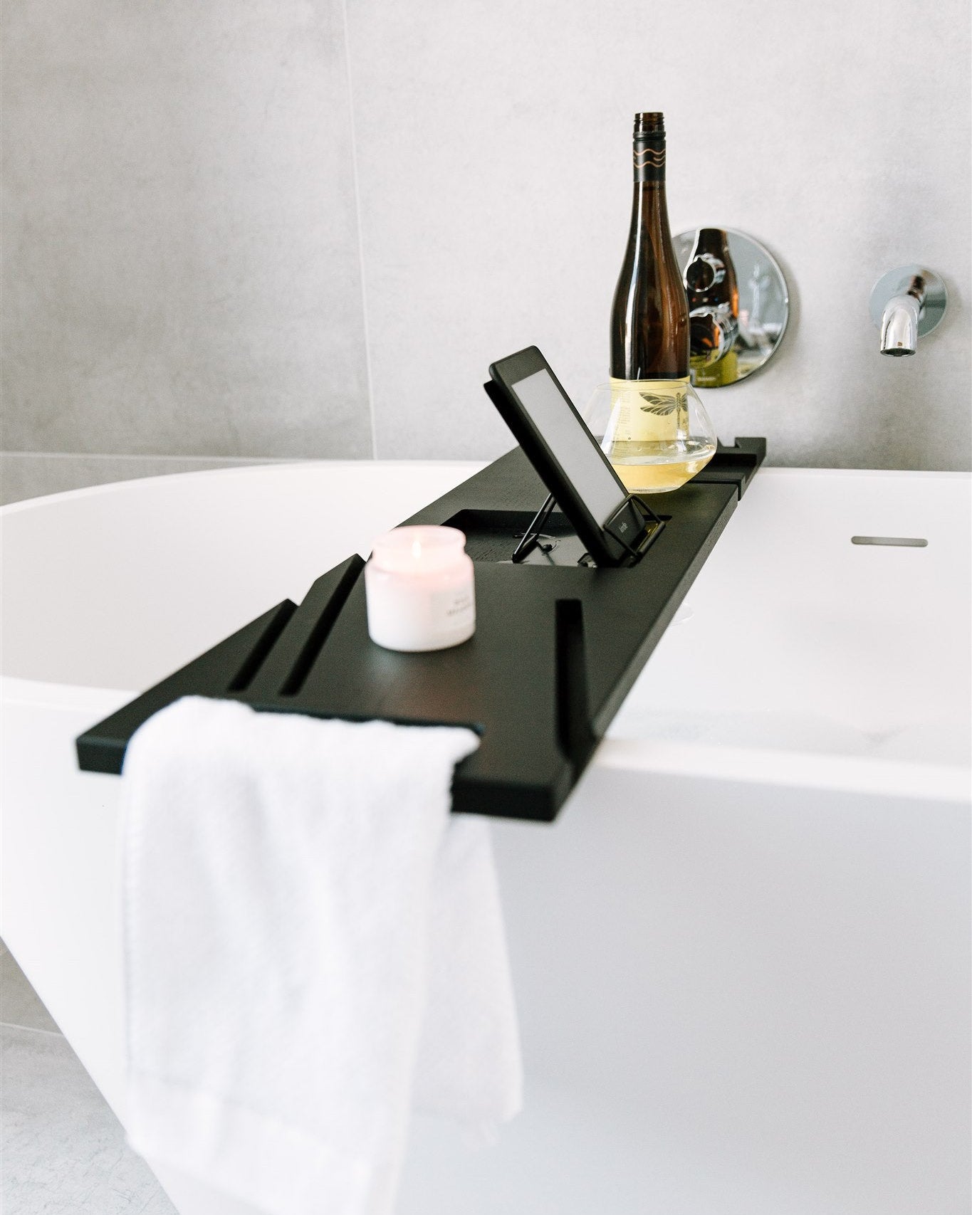 Black bathtub tray with a bottle, candle, and phone on a white bathtub.