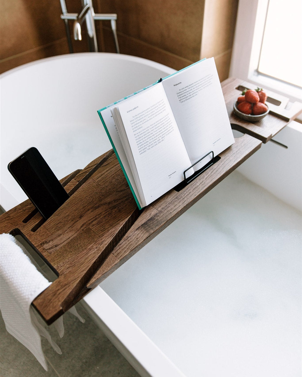 Wooden bathtub caddy with book and phone on a white bathtub
