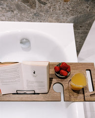 Wooden tray with a book, glass of juice, and bowl of strawberries on a bathtub.