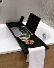 Black bathtub tray with laptop, glass, and bowl on a white bathtub.