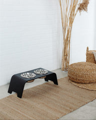 Black elevated pet feeder with two bowls on a textured rug next to a wicker chair and dried plants.