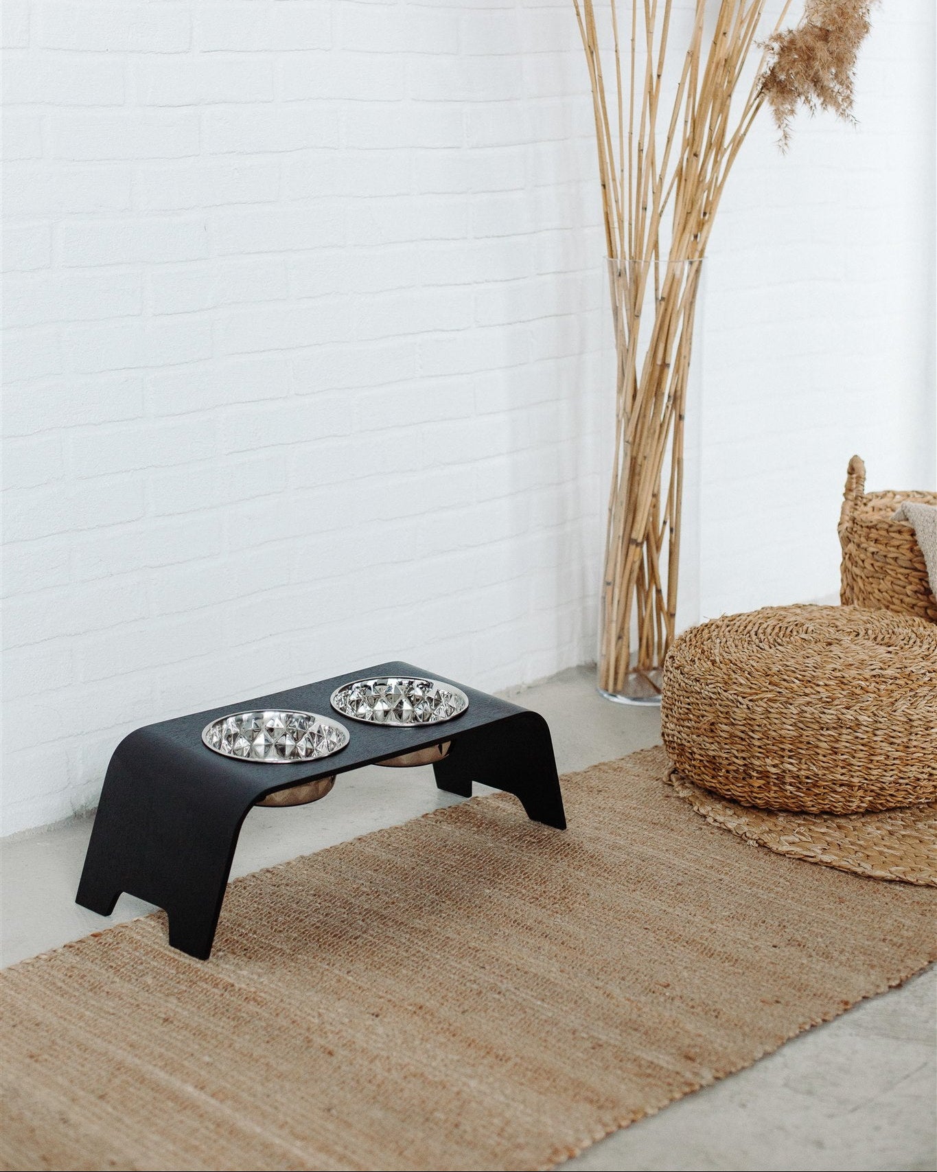 Black elevated pet feeder with two bowls on a textured rug next to a wicker chair and dried plants.