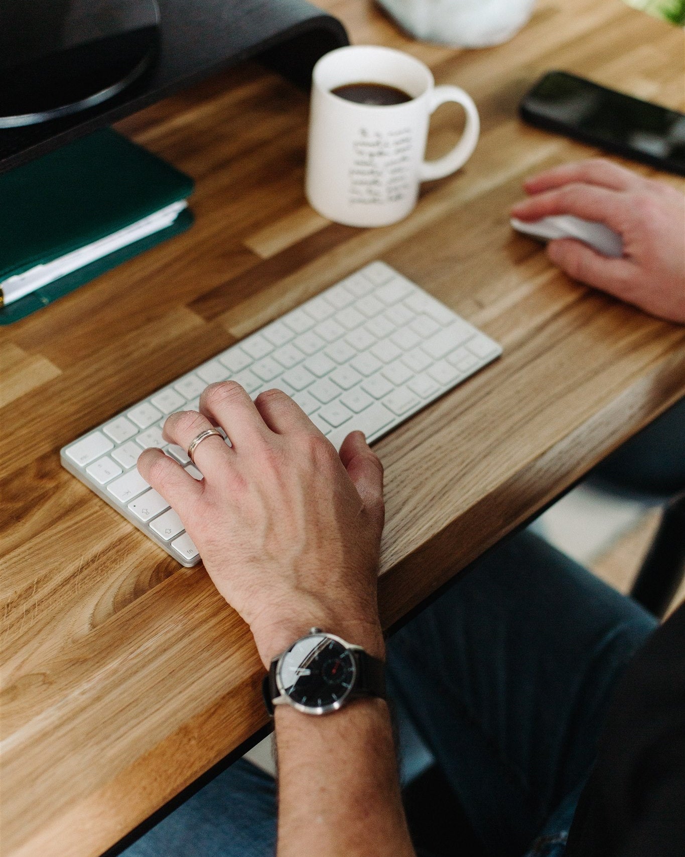 Person using a keyboard on a wooden desk with a mug and phone in the background