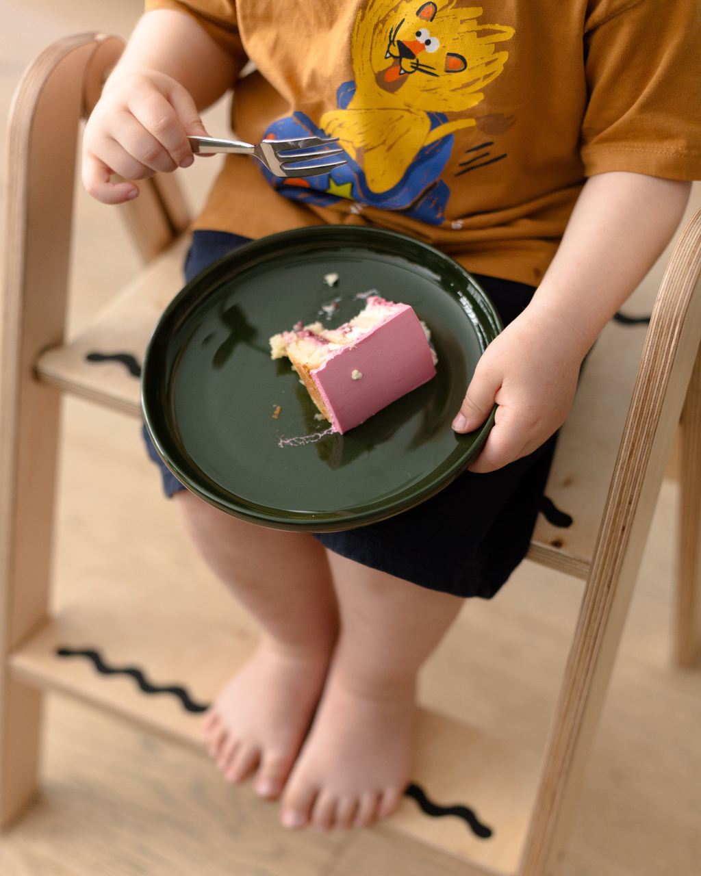 Child sitting on a high chair with a green plate holding a piece of cake.