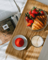 Wooden breakfast tray board with strawberries, croissant, and dessert on a light surface.