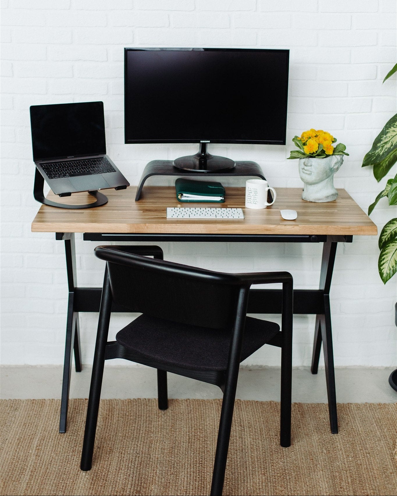 Home office setup with a wooden desk, laptop, monitor, and chair against a white brick wall.