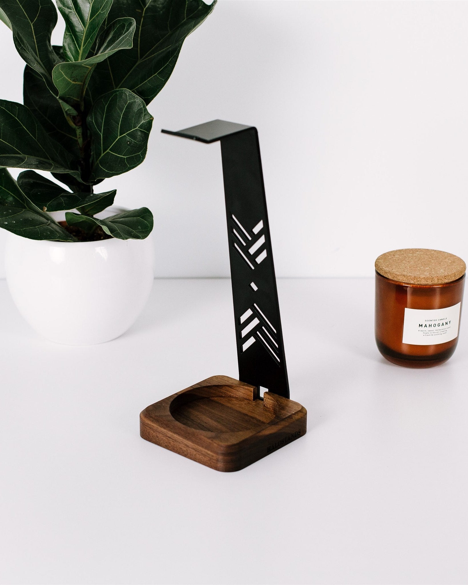 Black metal desk lamp with wooden base next to a candle on a white surface with a plant in the background