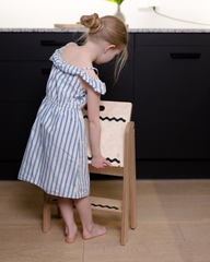 Child in a blue striped dress standing on a wooden stool in a kitchen.