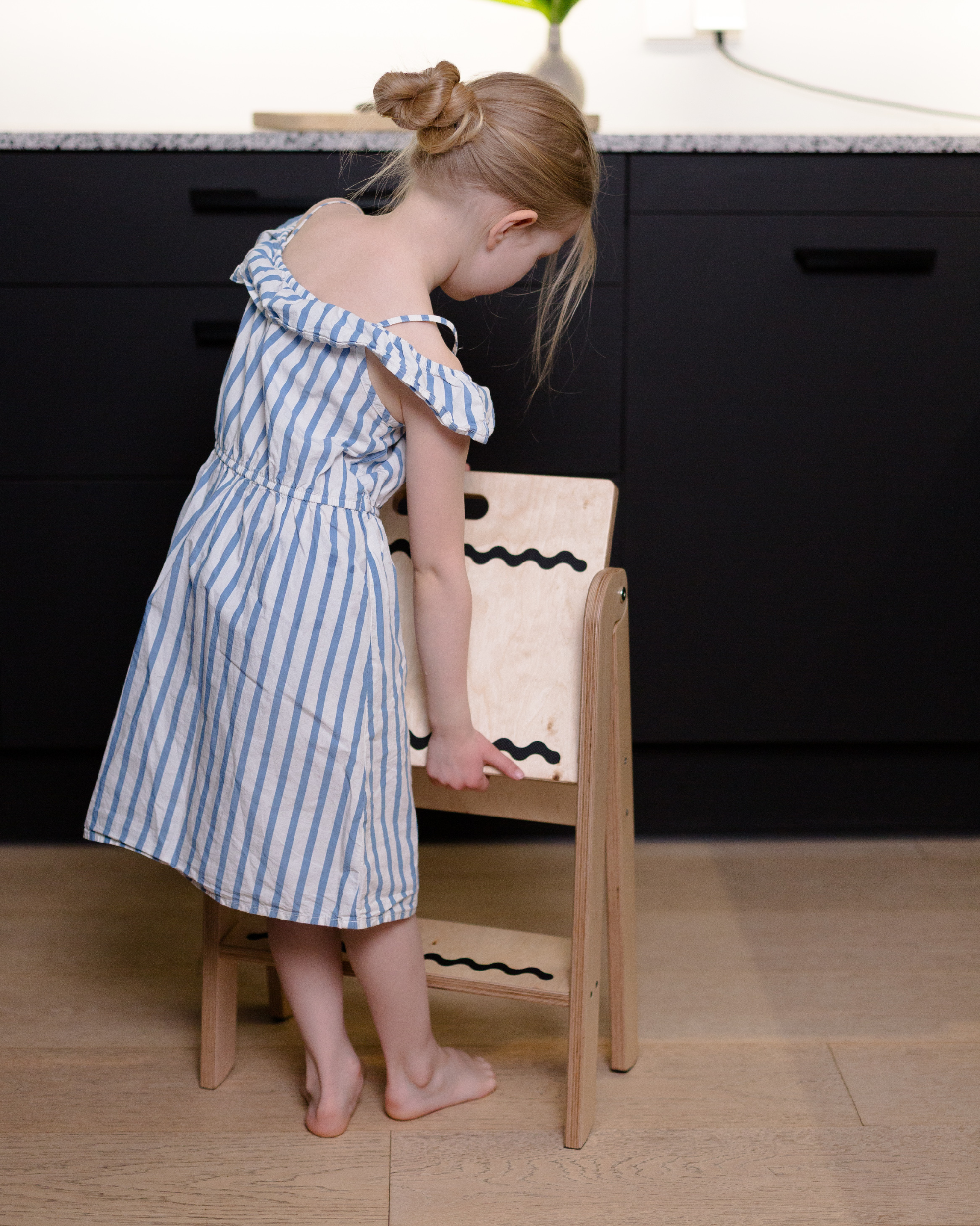 Child in a blue striped dress standing on a wooden stool in a kitchen.
