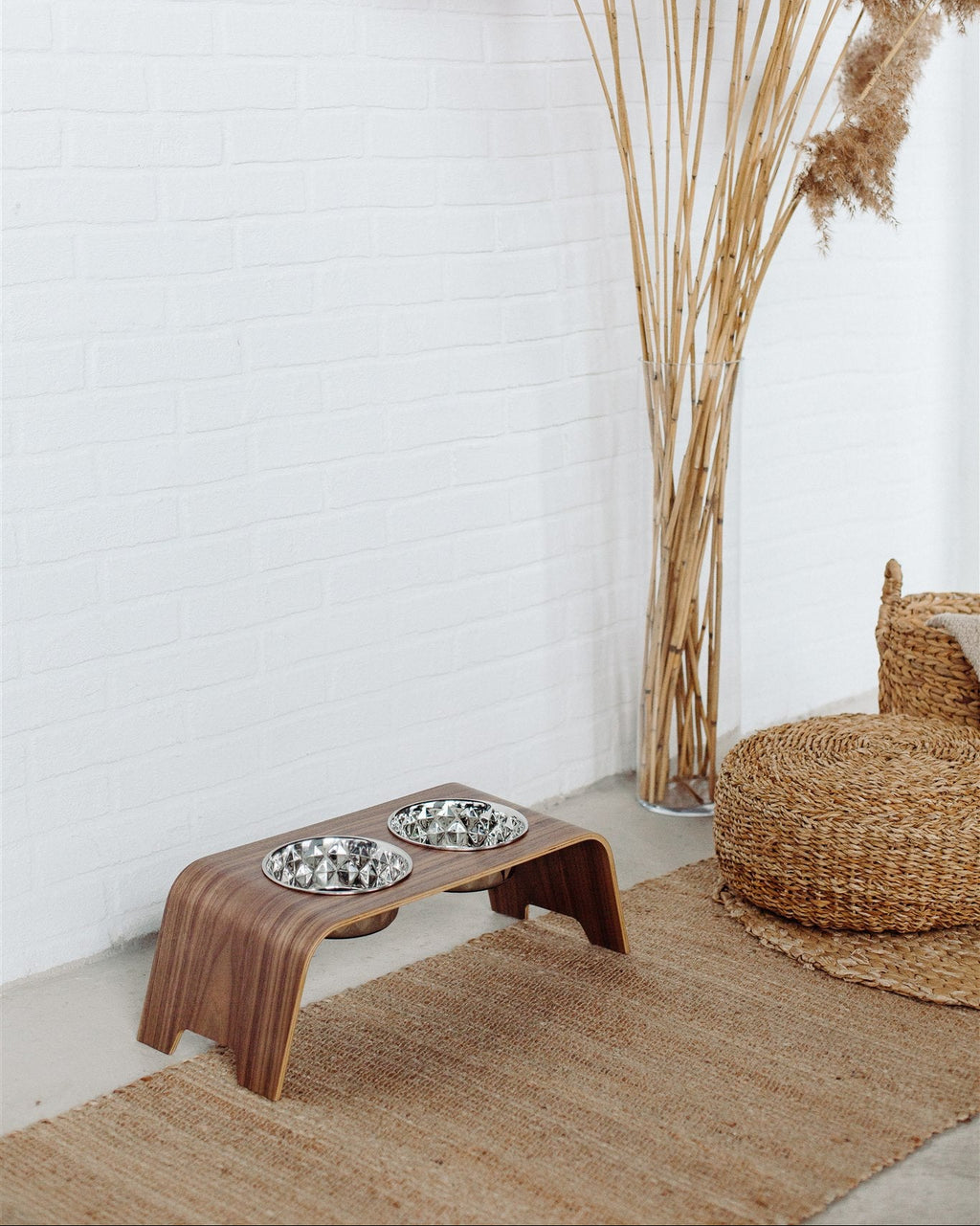 Wooden pet feeder with two bowls on a rug next to a wicker chair and tall pampas grass plant.