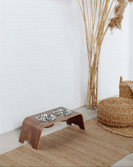 Wooden pet feeder with two bowls on a rug next to a wicker chair and tall pampas grass plant.
