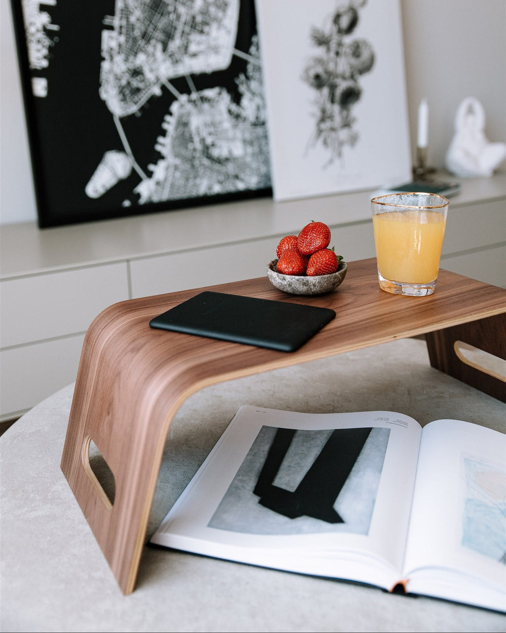 Small wooden table with a glass of orange juice, bowl of strawberries, and open book on a light-colored floor.